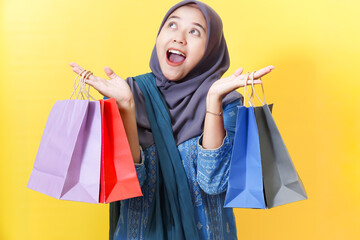 Excited woman in hijab holds shopping bags against yellow background.