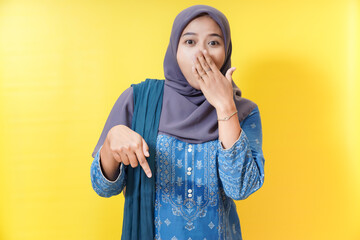 Surprised woman in hijab pointing down on yellow background.
