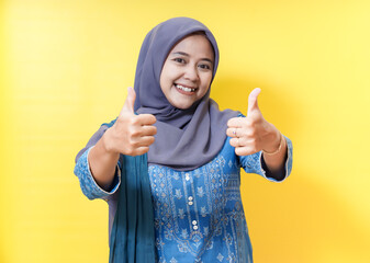 Smiling woman in hijab giving thumbs up on yellow background.
