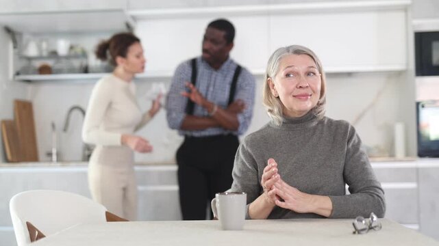  Elderly woman smiling during family quarrel between couple of adult man and woman in kitchen