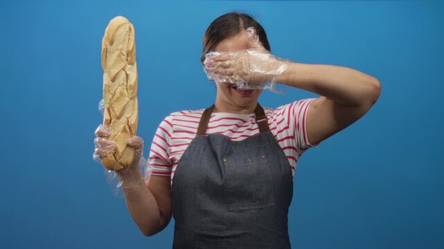 Woman holding a baguette and covering her eyes with a gloved hand in a blue studio; playful baking joy.