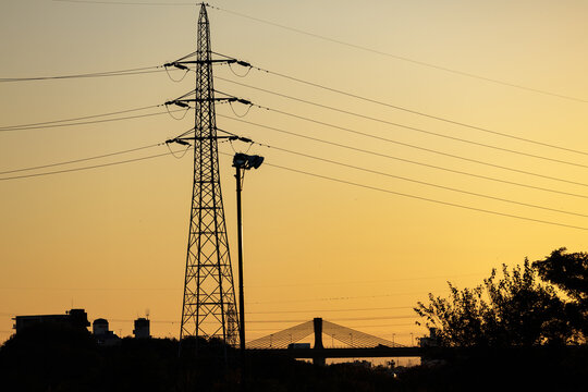 Golden sunset silhouettes landscape of high voltage tower and taut power lines towering above trucks crossing cable-stayed suspension bridge.