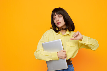 Fototapeta premium Young professional or student wearing a yellow shirt holds a laptop against a bright orange wall, showing frustration as they check the time and face a looming deadline.