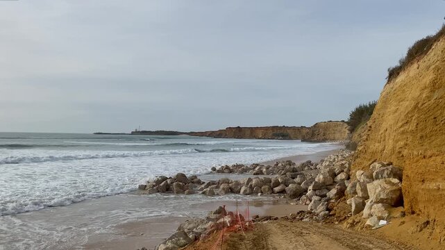 Sea defences of piled rocks protect the eroding cliffs while waves push onto the narrow winter beach at Fuente del Gallo