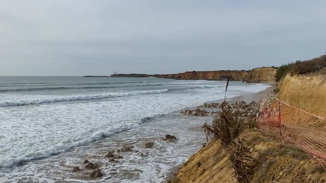 Sea defences of piled rocks protect the eroding cliffs while waves push onto the narrow winter beach at Fuente del Gallo