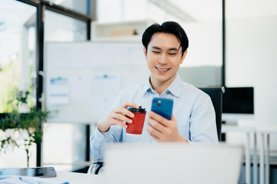 Confident Asian businessman typing laptop computer and digital tablet while holding coffee at office - Powered by Adobe