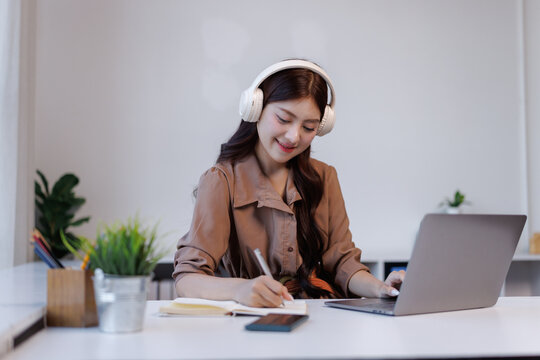 Young woman studying online, writing notes, wearing headphones - Powered by Adobe