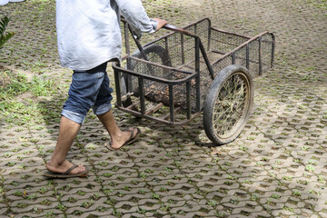 Asian worker pushing metal cart for manual labor on paved outdoor path. This diligent man is walking with his barrow, showing simple, everyday life and work