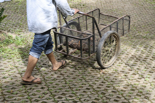 Asian worker pushing metal cart for manual labor on paved outdoor path. This diligent man is walking with his barrow, showing simple, everyday life and work