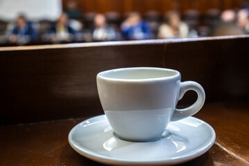 An empty coffee cup on a wooden desk with the blurred background of a plenary session in a government assembly, symbolizing a break or the long hours of political debate and decision-making.