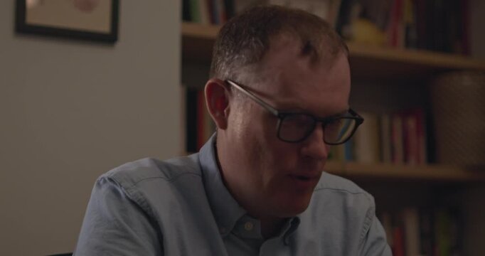 A close up of a man in glasses at work talking and laughing on a virtual meeting. Office library in the background.