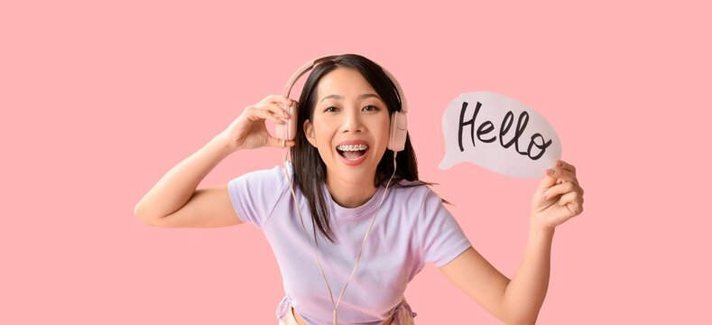 Young Asian woman in headphones holding speech bubble with word HELLO on pink background