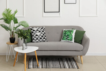 Cozy sofa with cushions and houseplants on tables near white wall