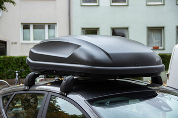 Black roof box mounted on a car, showcasing a sleek design and aerodynamic shape, parked in an urban environment with residential buildings in the background