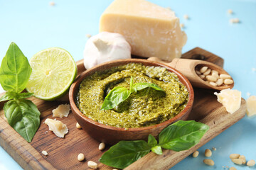 Wooden board with bowl of delicious pesto sauce and ingredients on blue background