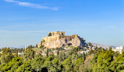 Ancient Acropolis hill above Athens upon bright day, viewed over green trees under clear blue sky
