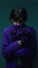 A woman in a purple sweater holds vibrant violet flowers, symbolizing tranquility and introspection against a deep green background.