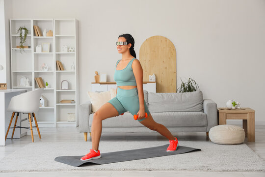 Sporty African-American woman in VR glasses training with dumbbells at home