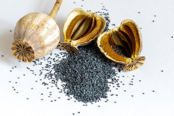 Close up of dried poppy seed pods with a mound of tiny gray seeds scattered on a clean white surface. Natural, rustic, minimal composition suitable for culinary, botanical, or art themes.