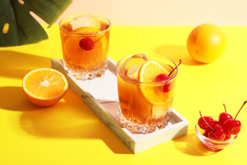 Glasses of tasty Old Fashioned cocktail with cherries and monstera leaf on yellow table against beige background