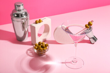 Composition with glass of tasty martini cocktail, green olives and shaker on pink background