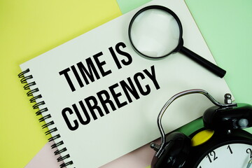 Time Is Currency text written on notepad with alarm clock and magnifying glass on pastel background