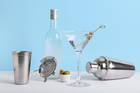 Composition with glass of tasty martini cocktail, green olives and bartender utensils on light table against color background