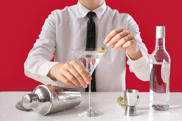 Female bartender making tasty martini cocktail at table against color background