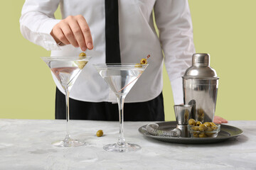 Female bartender making tasty martini cocktail at light table against color background