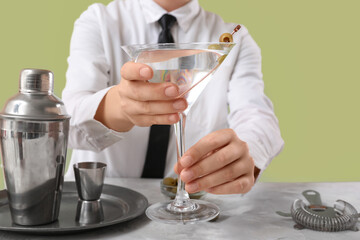 Female bartender with glass of tasty martini cocktail, green olives, shaker and strainer at table against color background