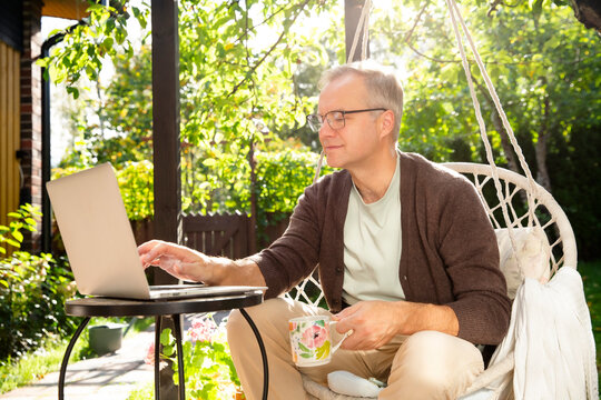 Middle-aged man working remotely from laptop and drinking coffee in hanging chair on sunny patio, relaxed lifestyle and modern home office. - Powered by Adobe
