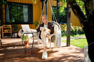 Middle-aged man working remotely from laptop and drinking coffee in hanging chair on sunny patio, relaxed lifestyle and modern home office.