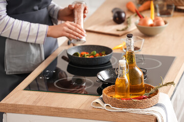 Bottles of sunflower oil with hot chili peppers and rosemary on table against woman cooking in kitchen, closeup