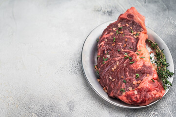 Uncooked strip loin new york beef steak with herbs and coarse salt on a silver plate beside vintage carving fork on soft gray textured background.