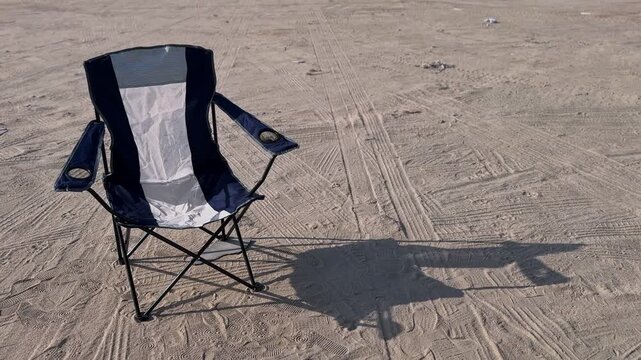 An empty blue and white folding camping chair sits alone on sandy ground covered in tire tracks and footprints. A solitary outdoor scene depicting a beach trip or desert camping adventure.