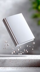 A blank white book is suspended in mid-air, with small droplets scattered around it, set against a textured background and a concrete surface.