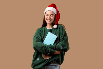 Happy young woman in Santa hat and Christmas sweater with book on beige background