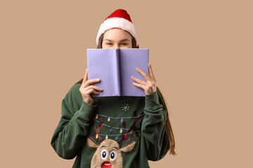 Happy young woman in Santa hat and Christmas sweater with book on beige background