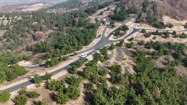 Aerial view of a white car driving on a mountain road. After crossing a three-way intersection, it turns left and continues deeper into the forest.