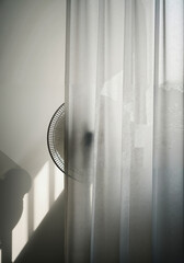 Standing fan partially visible behind sheer curtain in bright room  