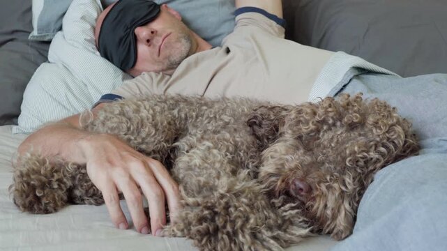 Maltipoo dog in bed with owner wearing a black sleep eye mask during a daytime nap, while the camera refocuses from the dog to the man. Funny pets and healthy lifestyle concept.