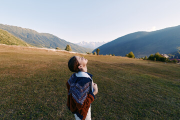 Fototapeta premium Girl with backpack standing in sunlit meadow looking up at distant mountains and valley, enjoying nature and rural landscape, freedom, exploration and a peaceful outdoor moment.