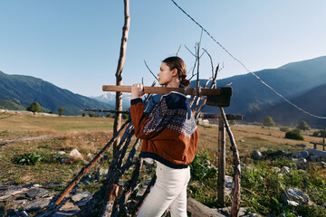 Fototapeta premium Woman carrying wood on rustic fence in mountains, wearing a warm sweater in a rural landscape; portrait of outdoor activity and traditional lifestyle on a sunny day.