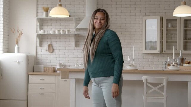 Positive plus size African American woman satisfied with appearance and body shapes, playfully slapping her buttocks, being happy and excited after weight loss success at home.