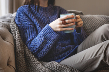 Female hands holding blue mug while sitting in oversized knitted sweater and warm blanket. Cozy autumn or winter atmosphere indoors with comfort
