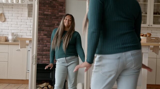 Portrait of positive plus size African American woman enjoying look and body shapes in mirror after successful weight program and dancing , expressing carefree mood and excitement in domestic room.