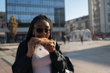 Beautiful black woman with sunglasses eating a slice of pizza on the city street on a sunny day