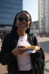 Beautiful black woman with sunglasses eating a slice of pizza on the city street on a sunny day