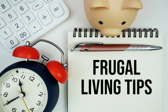 Frugal Living Tips text written notepad with calculator, piggy bank and alarm clock on white background