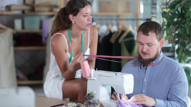 Cheerful young female tailor sitting on work table and playfully teasing male coworker working at sewing machine, making him look shy and slightly confused in workshop